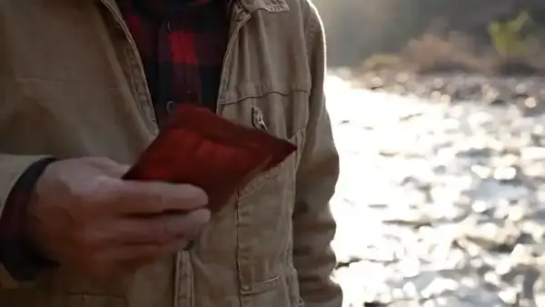 Weathered fingers sliding a small dark-red pouch into the breast pocket of a flannel shirt by the river